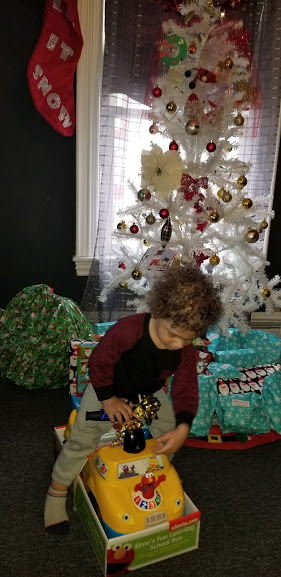 A child riding a small toy car in front of a white Christmas tree