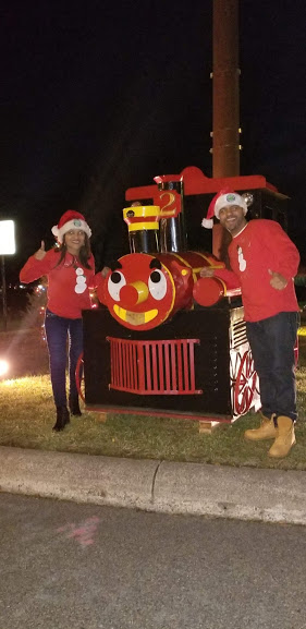 A woman and a man wearing red shirts and Santa hats doing thumbs up beside a red train attraction