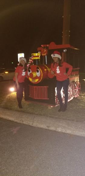 Two women wearing red shirts and Santa hats and a red train attraction, different angle