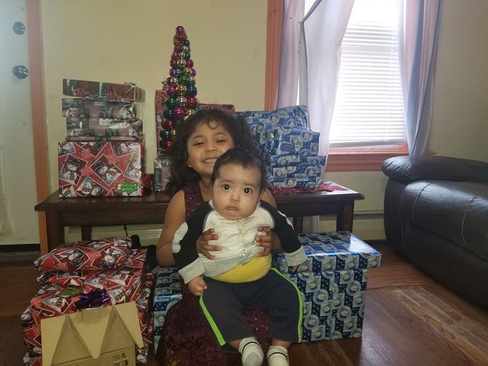 A little girl with pig tails carrying her younger sibling and posing with plenty of Christmas gifts behind them