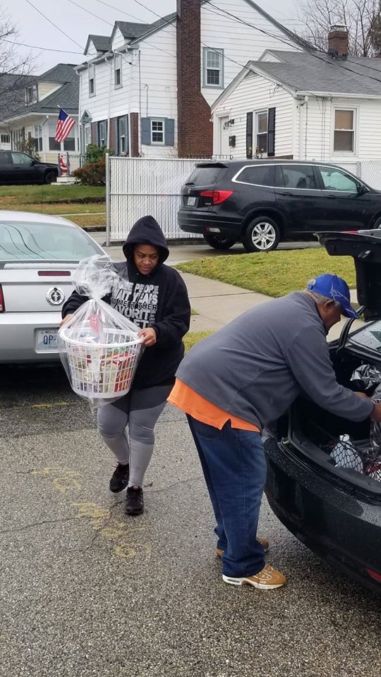 A woman in a hoodie carrying a basket of groceries and a man getting baskets from a trunk of a car