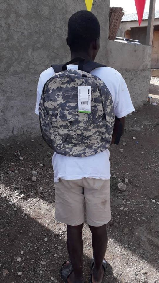 A boy in white shirt facing back and wearing a gray camouflage backpack