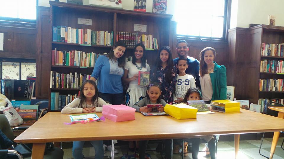 Four staff and two older children standing, and three young girls sitting at a table with books (slightly farther view)