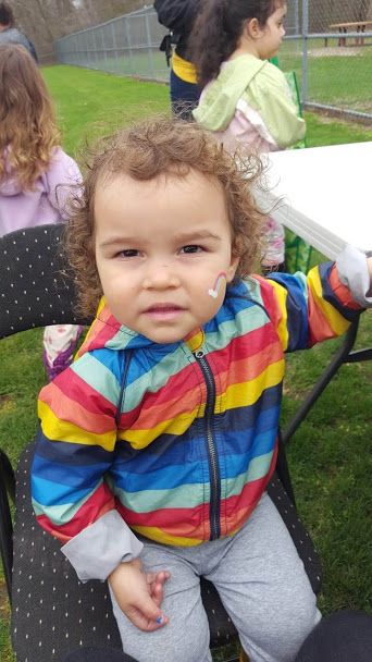 A child in rainbow jacket with a rainbow face paint, sitting