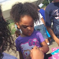 A little girl wearing a purple Hello Kitty shirt having her hand painted with a design