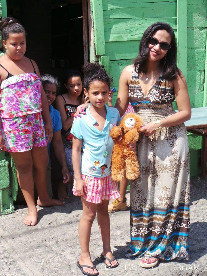 A woman in a dress giving a stuffed toy to a girl; other people behind them