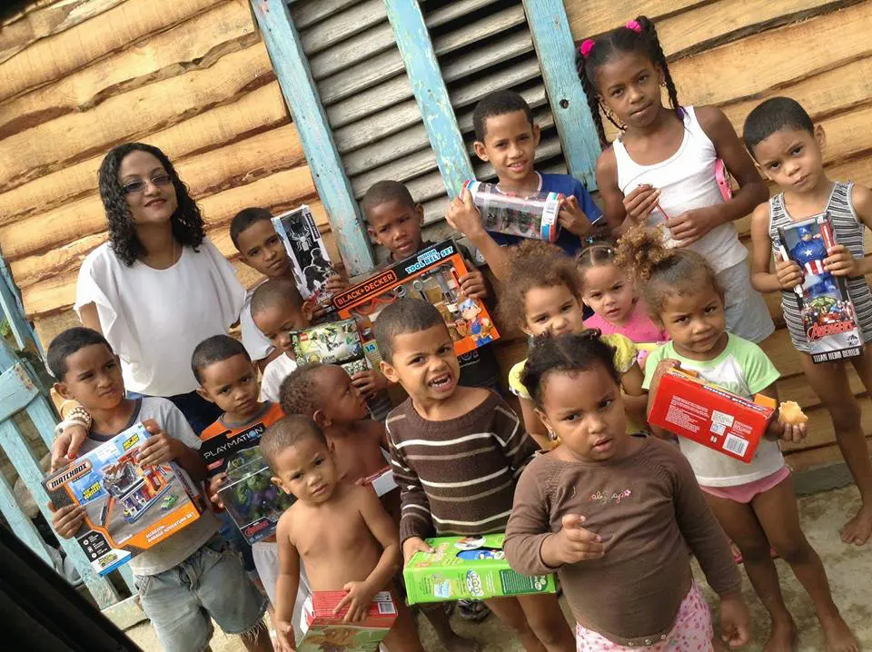 A staff and a big group of young children holding toys outside a wooden house