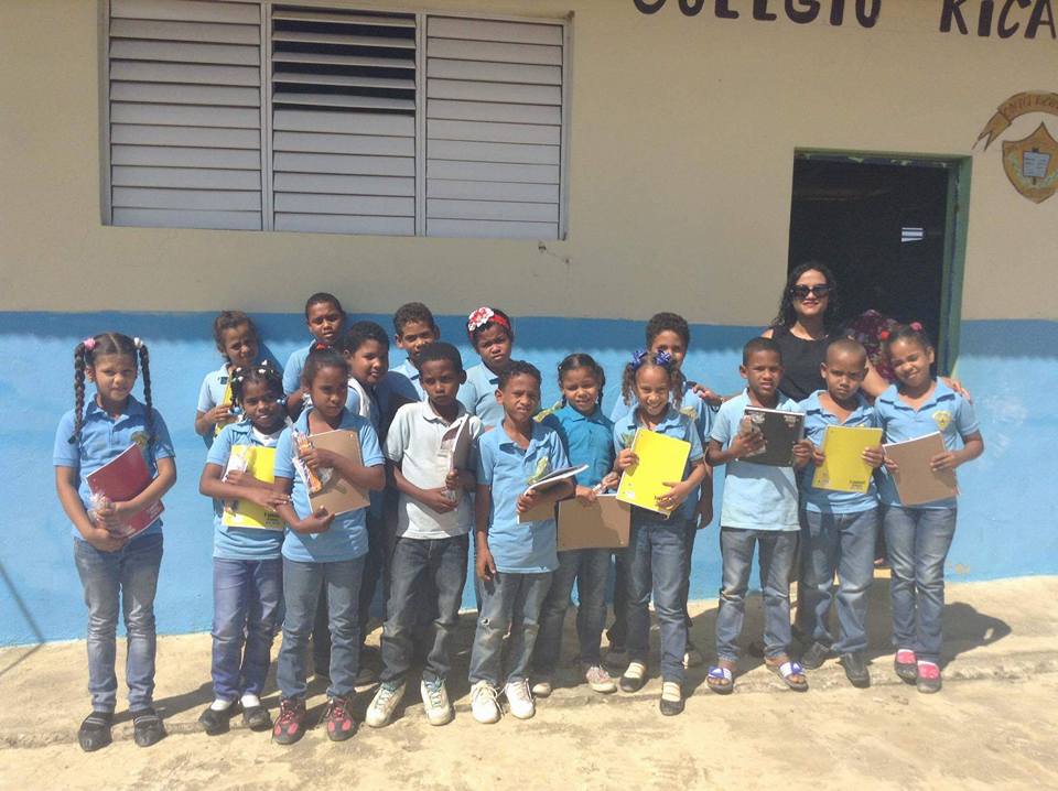 A group of children wearing blue polo shirt holding school supplies outside a classroom