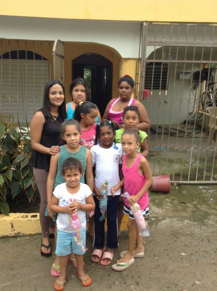 Our staff and a group of girls holding dolls in front of a yellow house