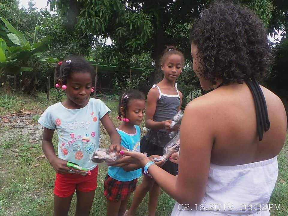 A woman in white dress giving toys to three girls on a grassy area