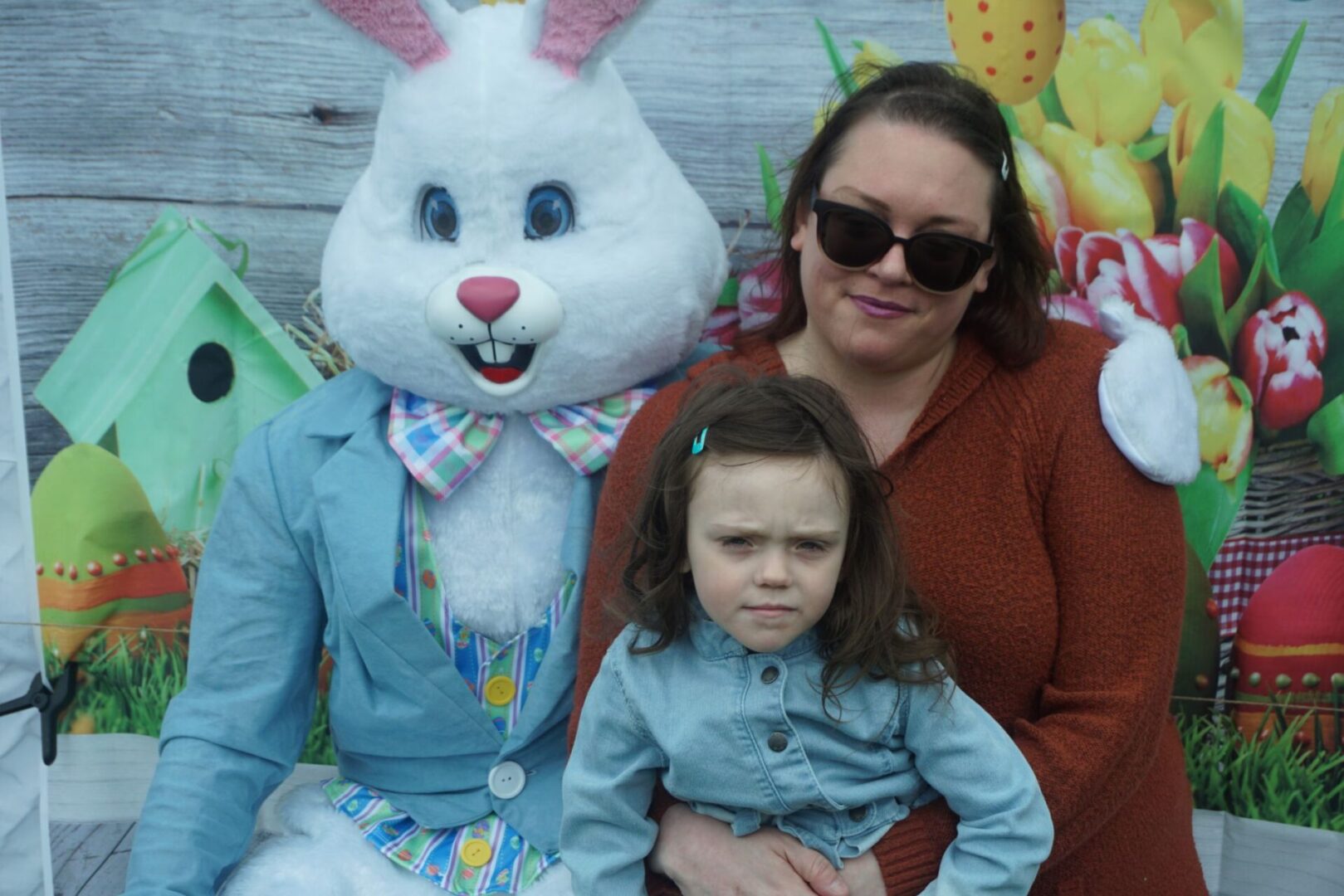 The bunny mascot with its arm around a mother wearing sunglasses and holding her daughter
