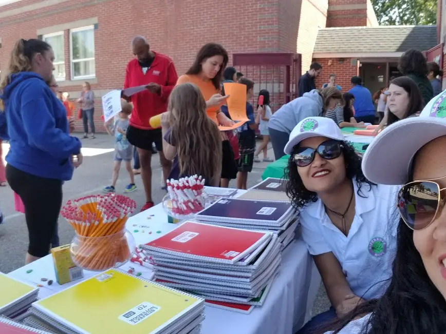 Two female staff wearing sunglasses and caps, smiling, and parents and children walking in front of their table