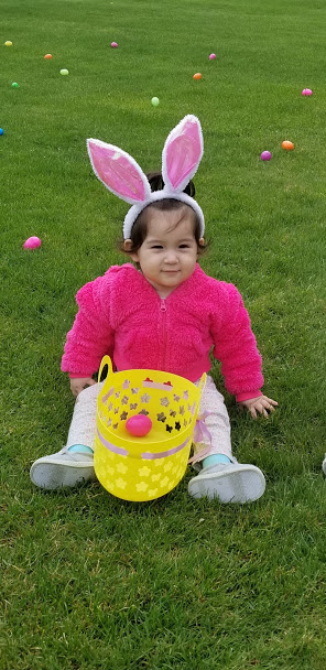 A baby in a pink coat and bunny ears sitting on the grass with her basket, smiling