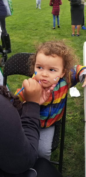 A young child wearing a rainbow jacket being face painted