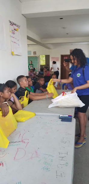 Our staff giving yellow tote bags to the children sitting at the long table, 3