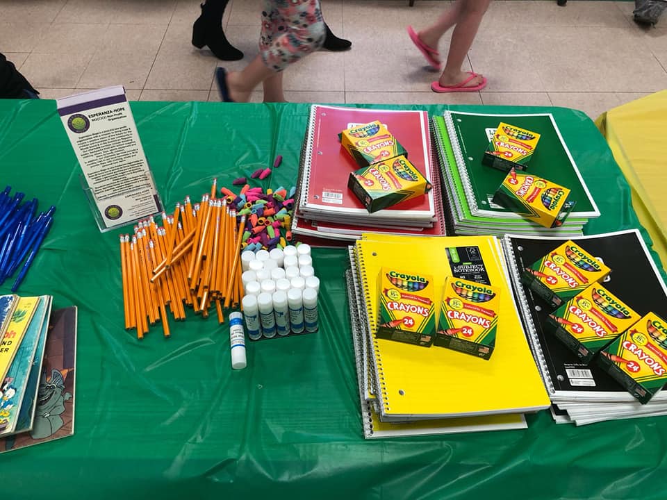 A table of notebooks, pencils, glue sticks, and other school supplies; people passing by
