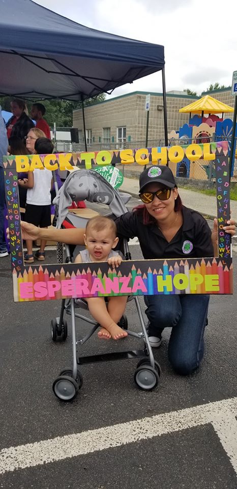 A baby in a stroller and a female staff in black polo shirt holding a colorful “Back to School” frame