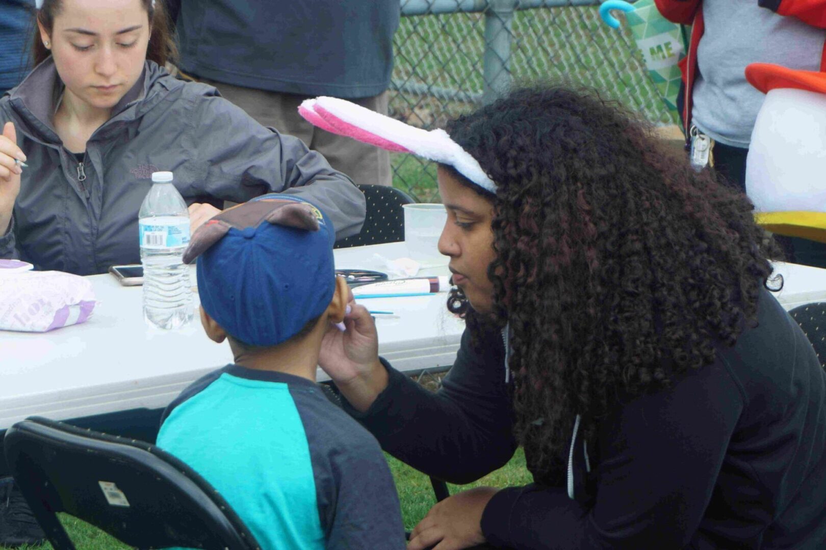 A woman in bunny ears face painting a boy with a cap, different angle