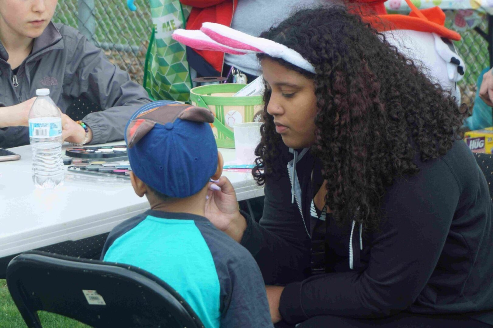 A woman in bunny ears face painting a boy with a cap
