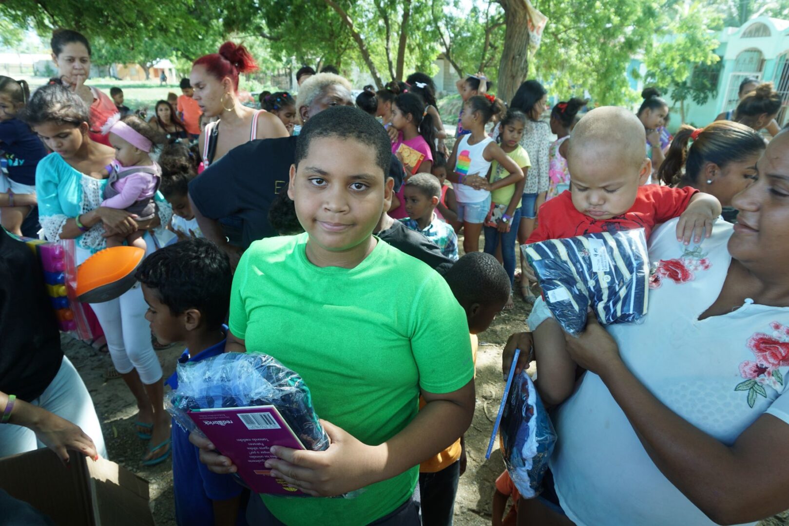 A girl in a green shirt holding a plastic of clothes and a book and beside her a mother and a baby