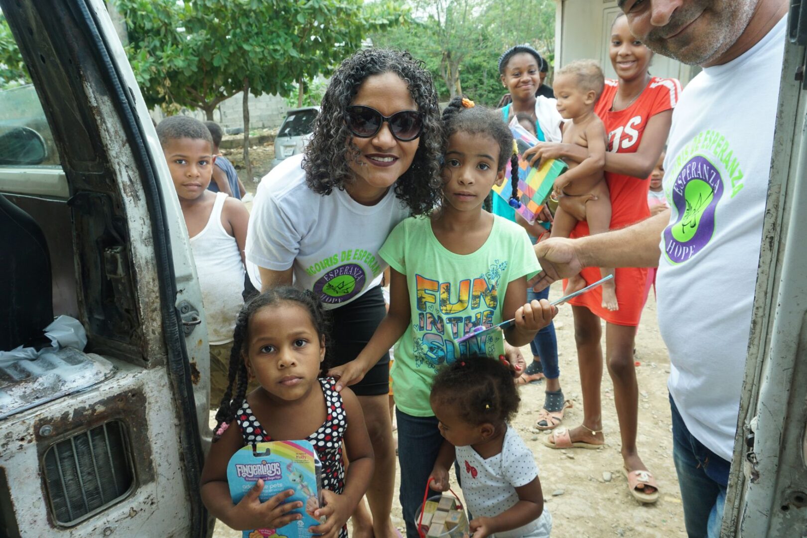 A staff and three girls holding toys outside; other children and parents at the back