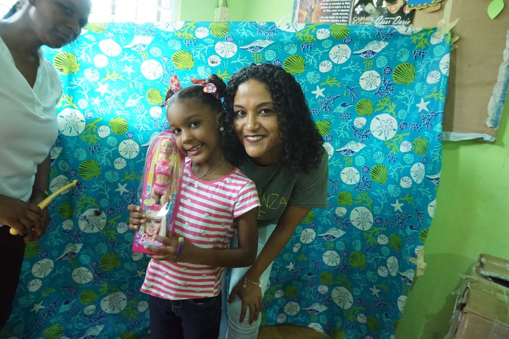 A staff and a girl holding a doll, smiling, standing against the seashell curtain