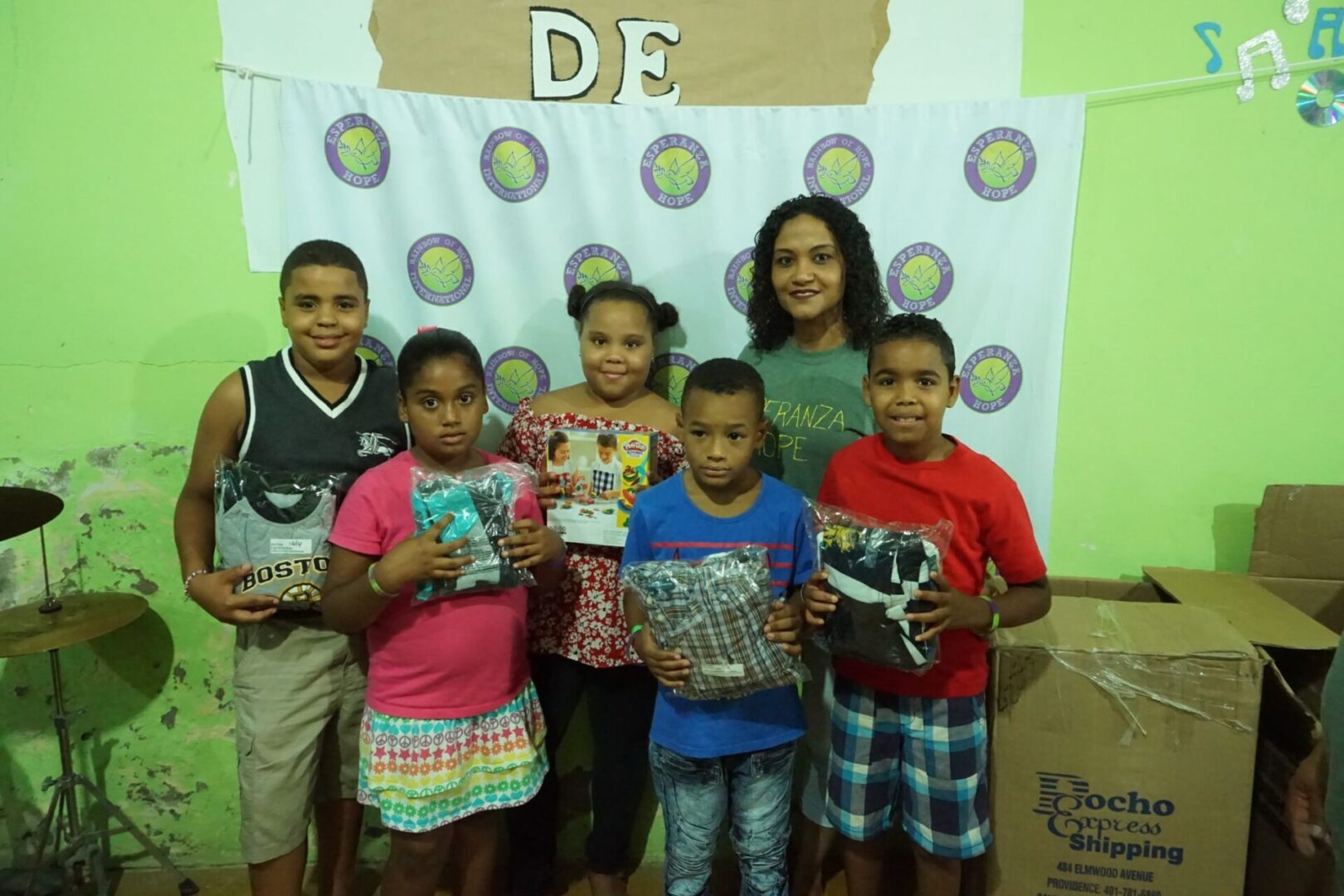A staff and a group of children holding packs of clothes, standing against a cloth with our logo