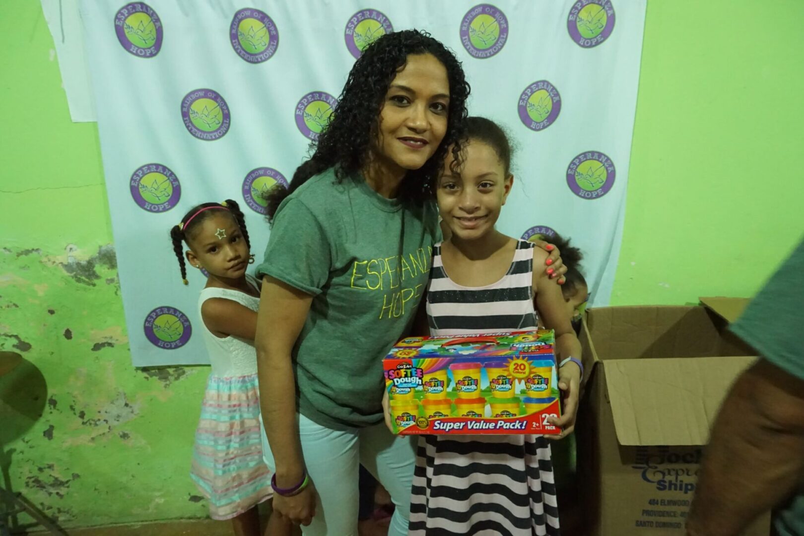 A female staff with her arms around a girl holding a box of toy; a little girl behind them
