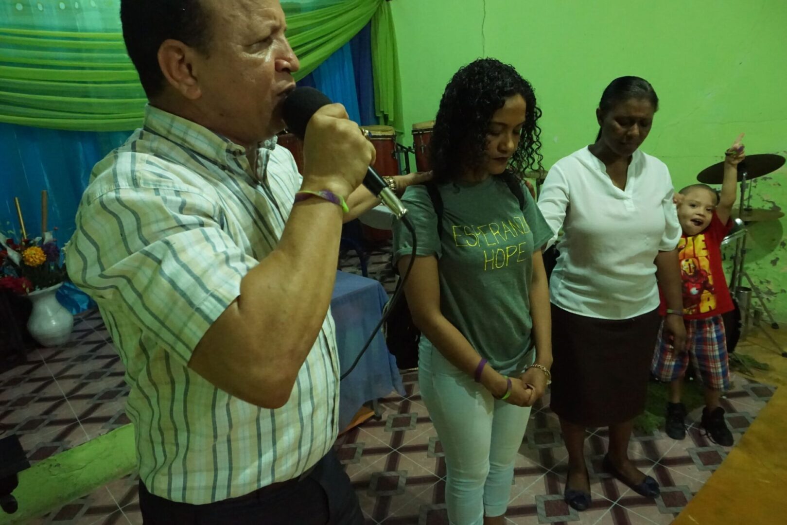 A man speaking into a mic and two women, all of them with their eyes closed; a child with his hands up