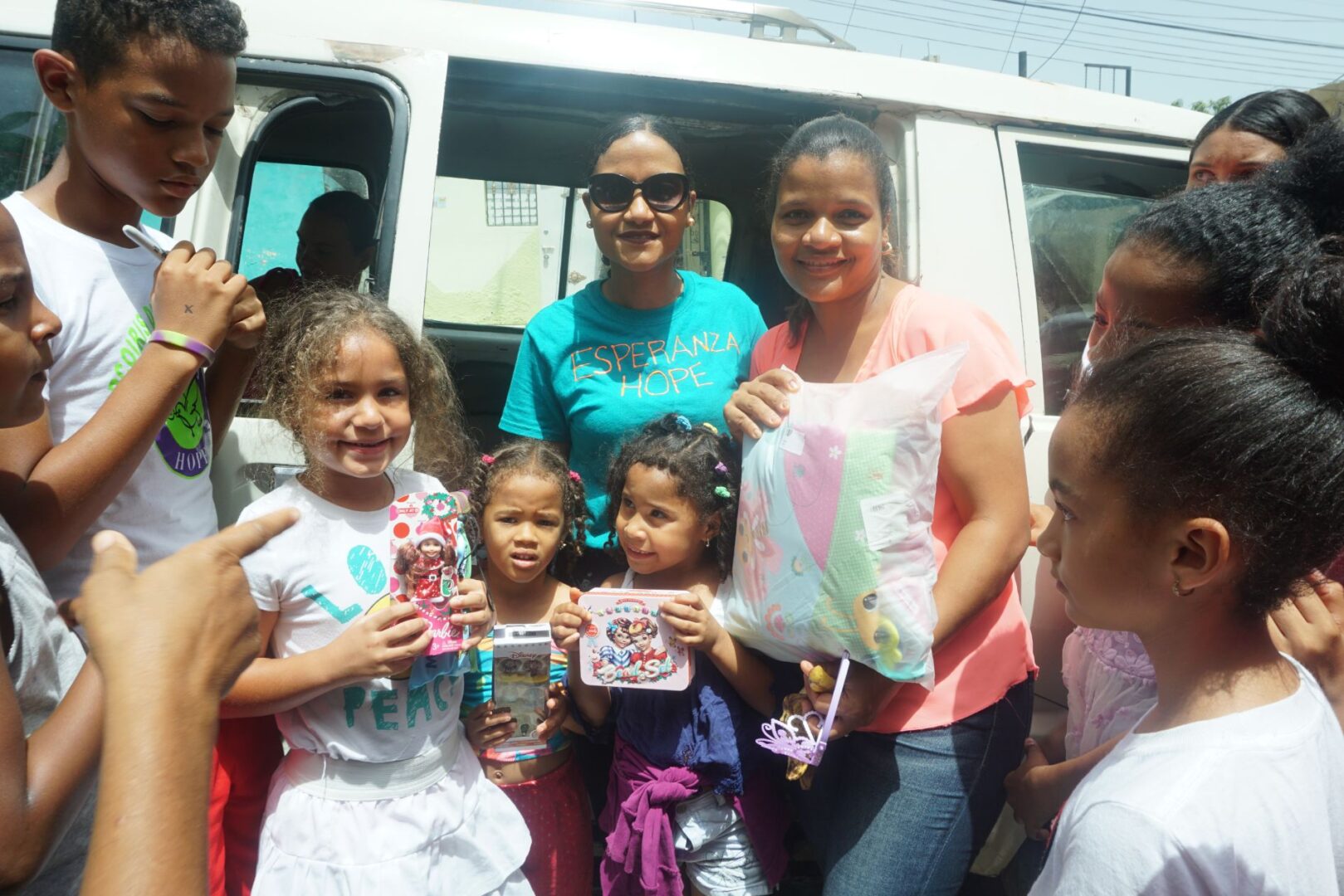 A staff in blue shirt, a woman, and a group of young girls holding toys in front of a vehicle with an opened door