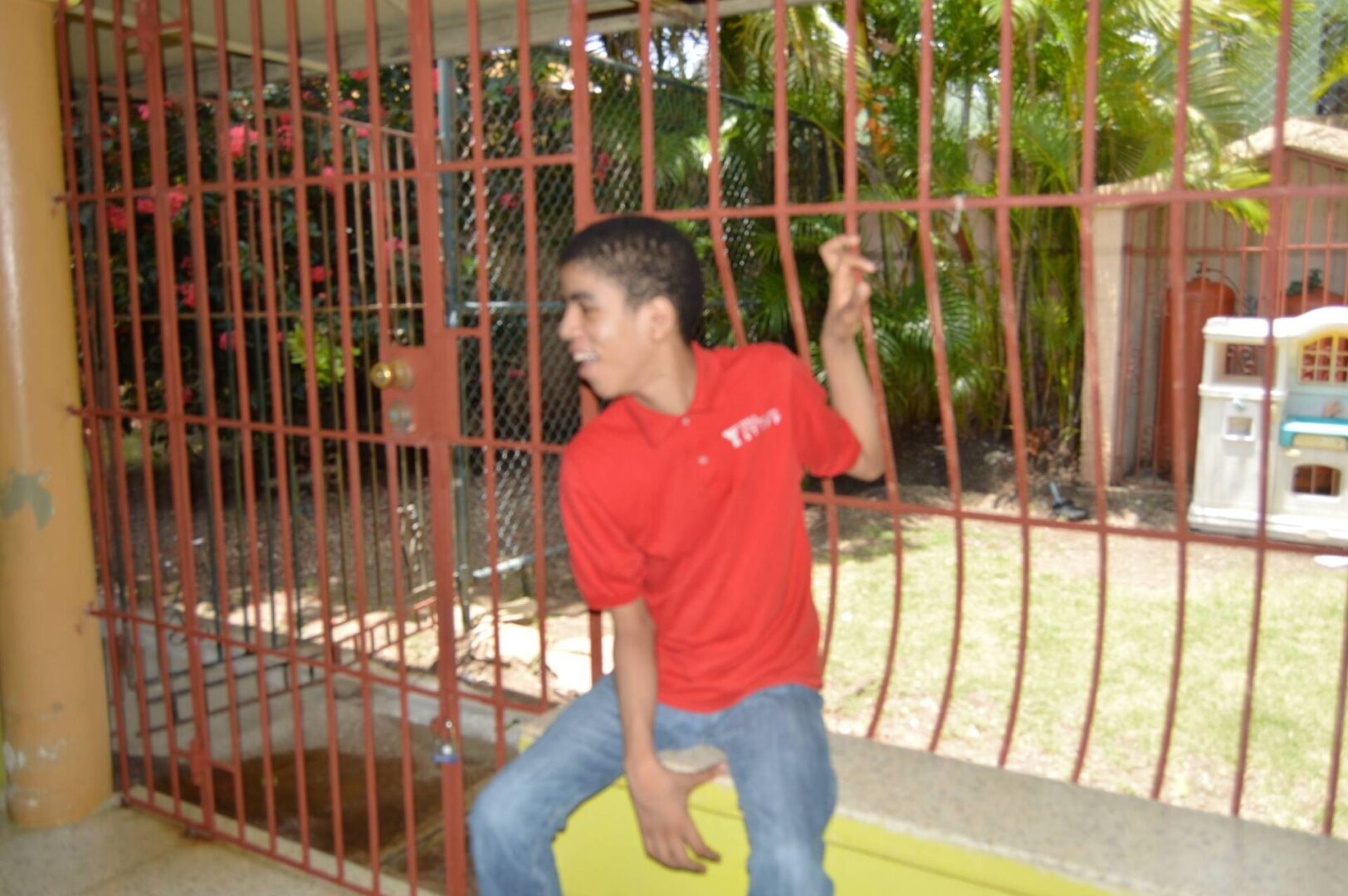 A boy in a red shirt sitting near the window bars, smiling
