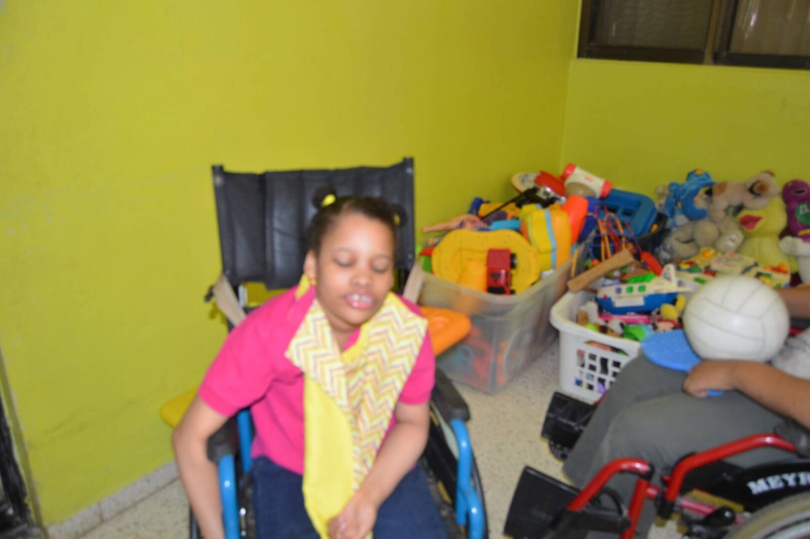 A girl in a wheelchair wearing a yellow zigzag cloth over a pink shirt