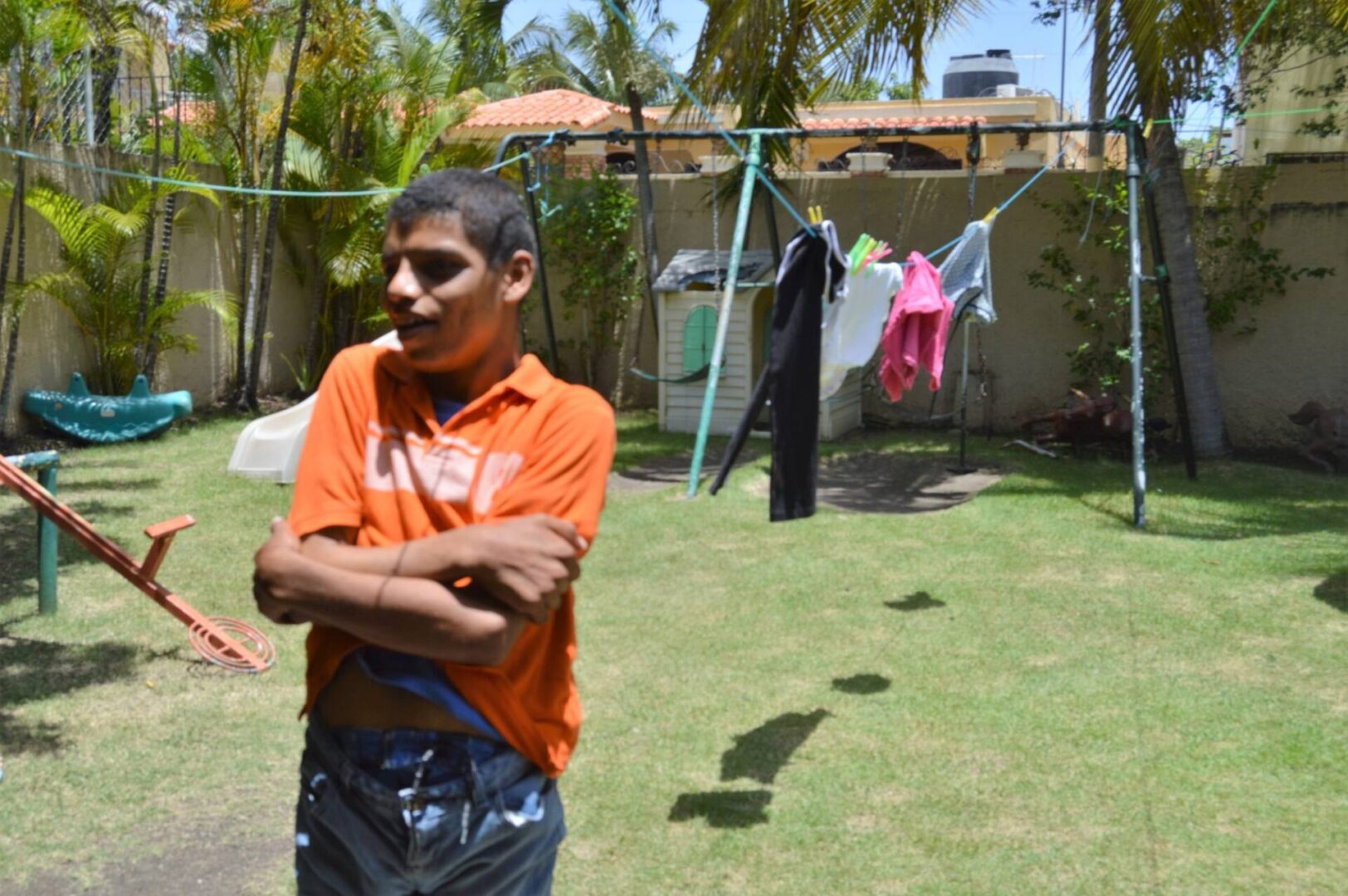 A boy in an orange shirt hugging himself and a clothesline and playground at his back