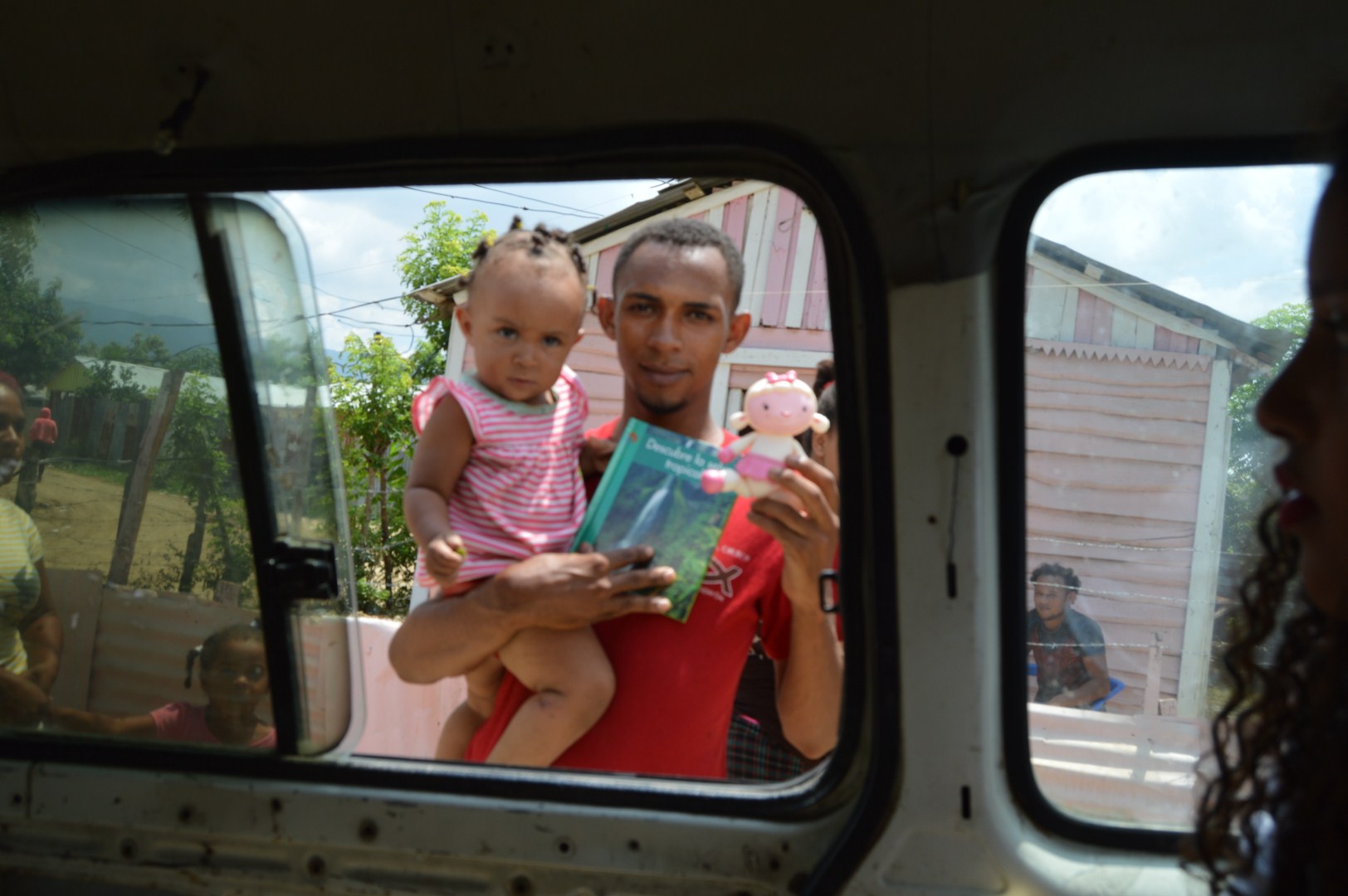 A man carrying a little girl holding a book and a toy outside the car’s window