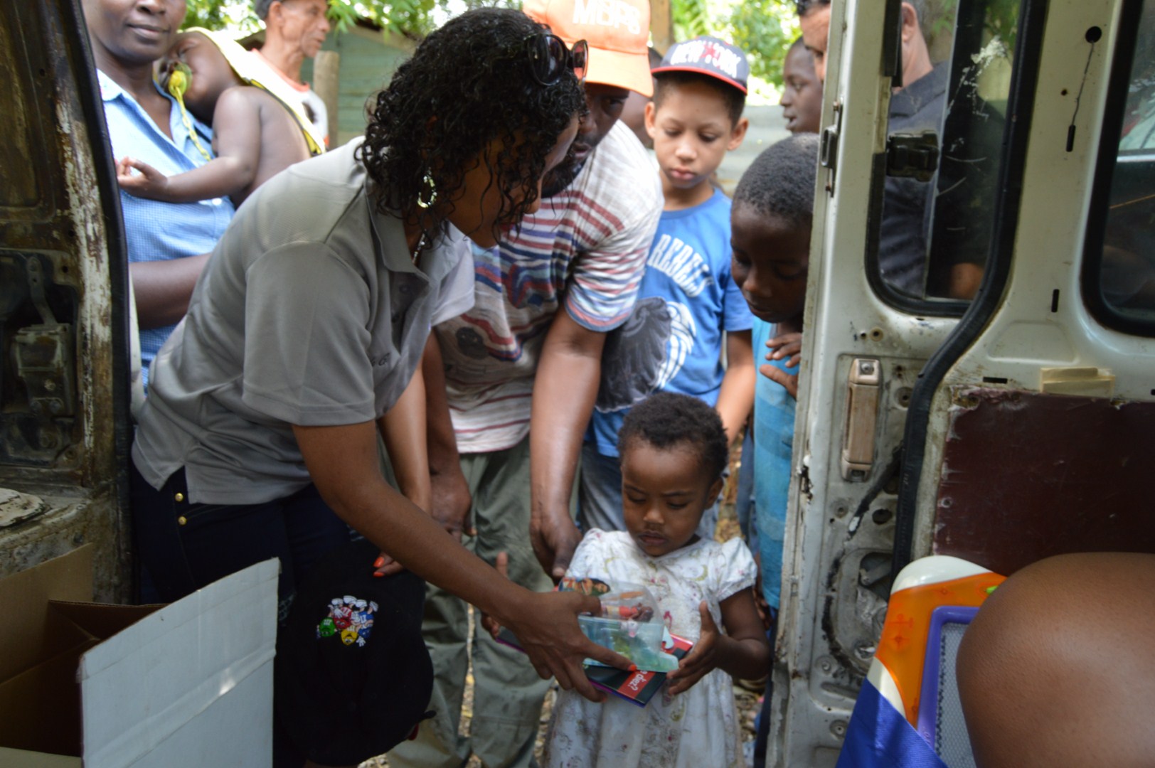 Our staff giving a toy to a little girl in a dress outside the car; children in line behind her