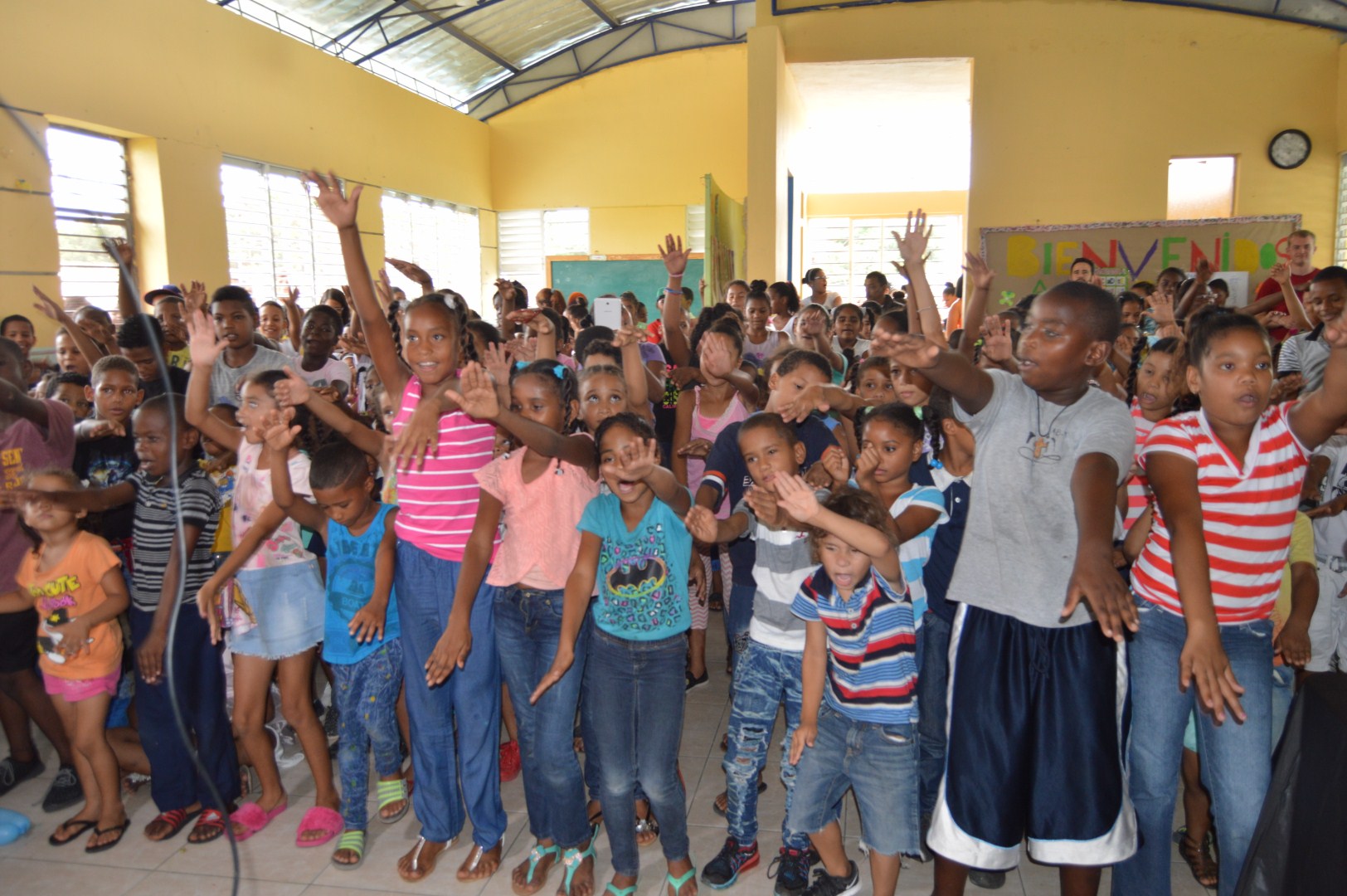 Children in a classroom, standing closely together and raising their hands