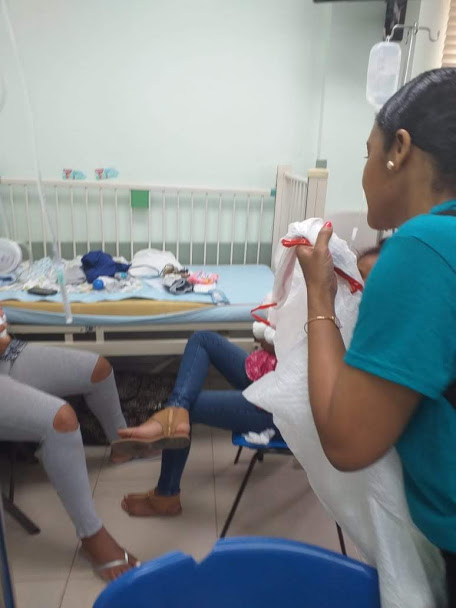 A woman in blue shirt getting something from a white plastic bag; someone in a hospital bed