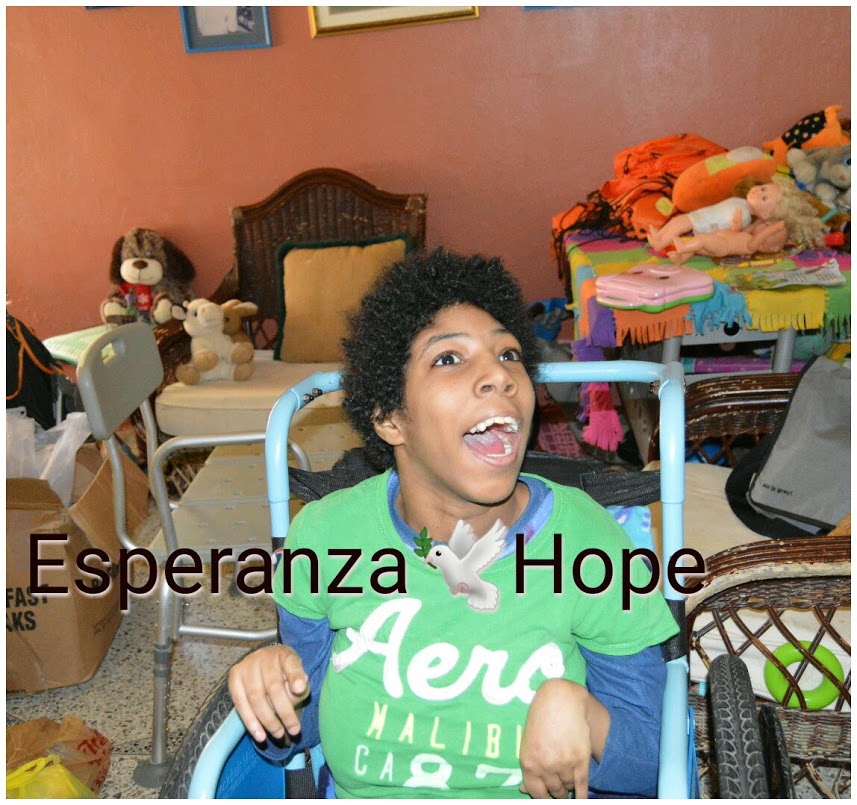 A girl on a blue wheelchair, smiling, and toys at the back. Text: Esperanza-Hope