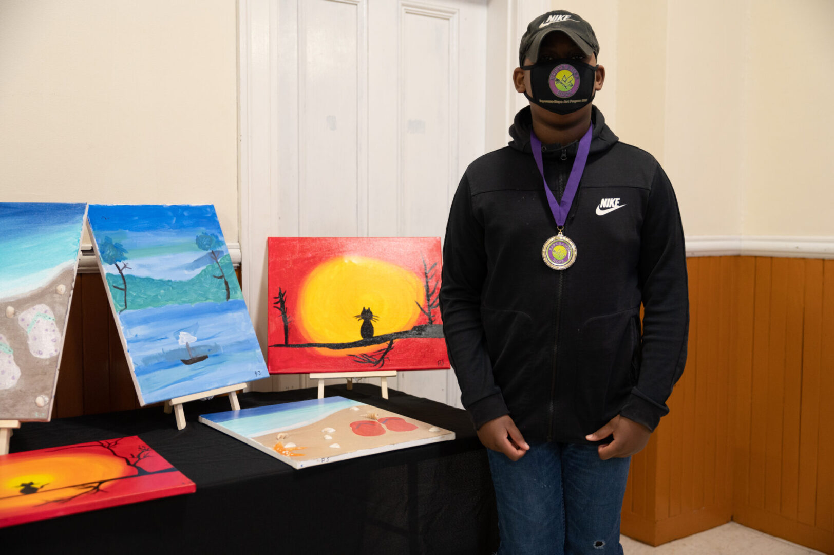 A boy in a jacket and wearing a medal, standing at the table with paintings