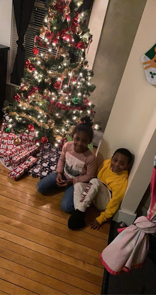 A boy and a girl sitting on the floor in front of a Christmas tree full of gifts