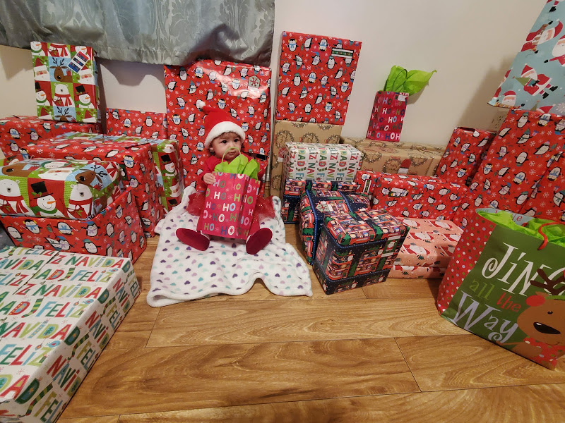 A cute baby wearing a Santa hat holding a gift and has plenty of Christmas gifts around her
