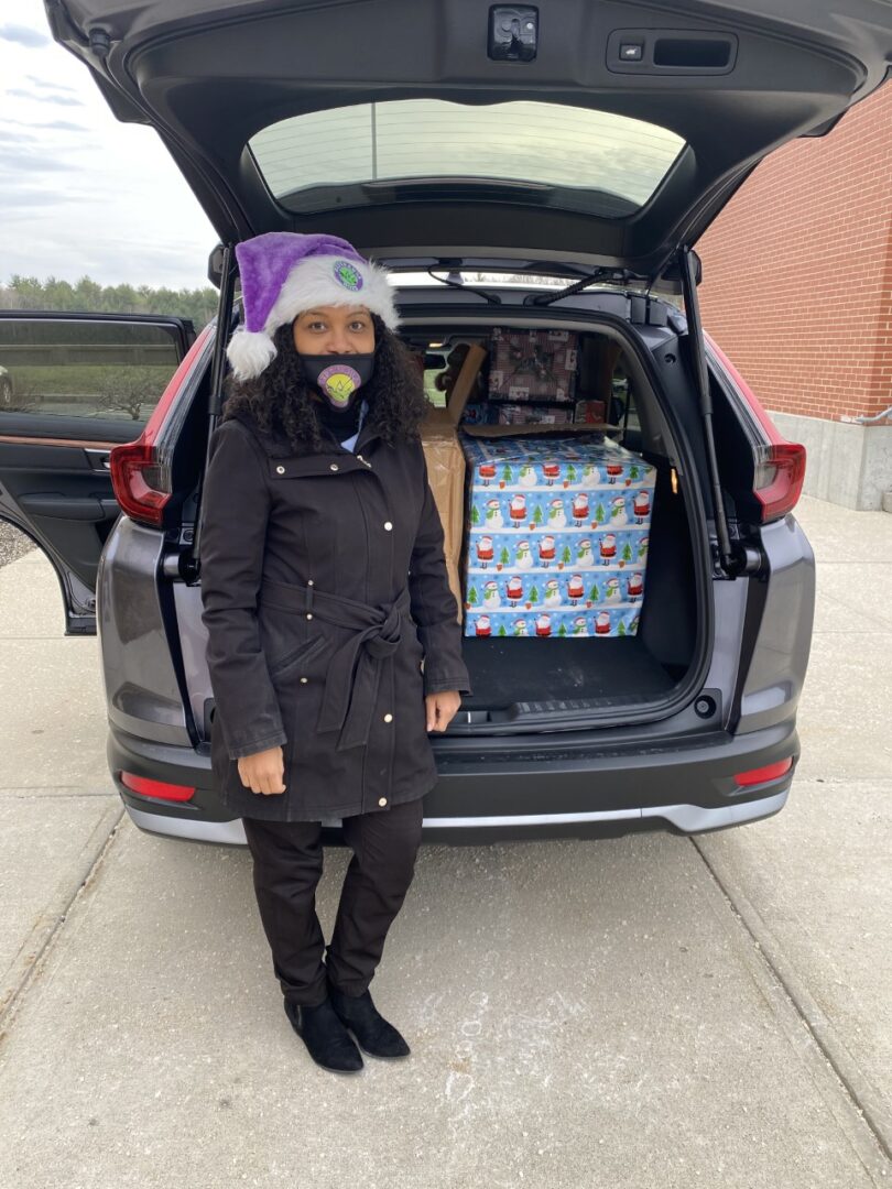 A woman wearing a mask and a purple Santa hat standing in front of an open car trunk with gifts