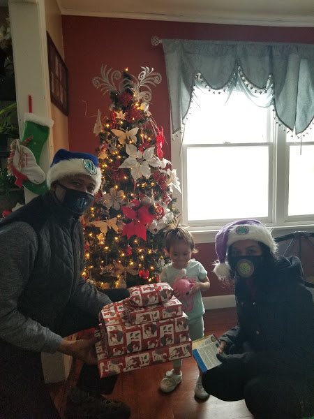 A man and a woman wearing Santa hats and carrying gifts and a little girl in front of a Christmas tree
