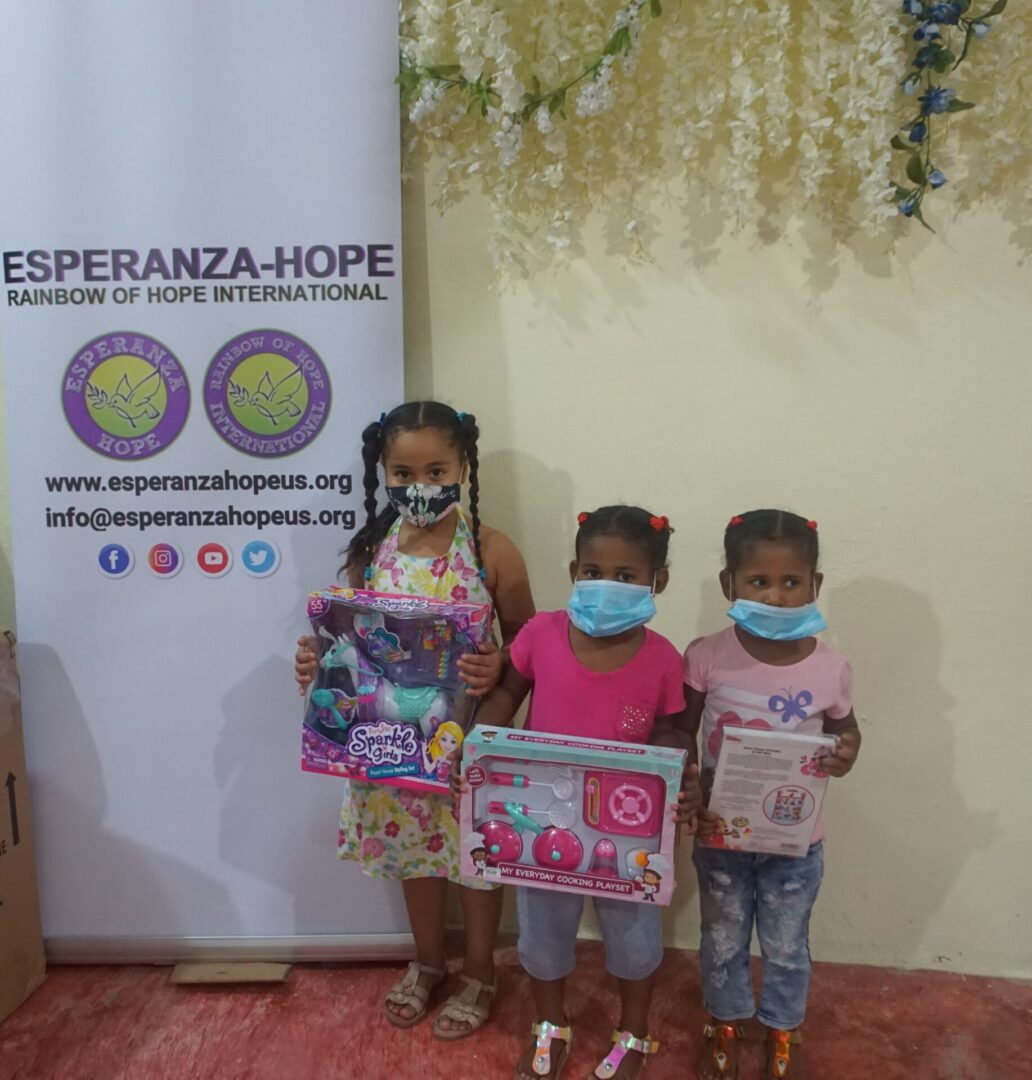 Three girls holding their toys beside an Esperanza-Hope tarpaulin