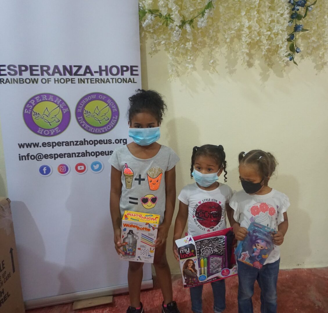 Three young girls holding toys beside the Esperanza-Hope tarpaulin