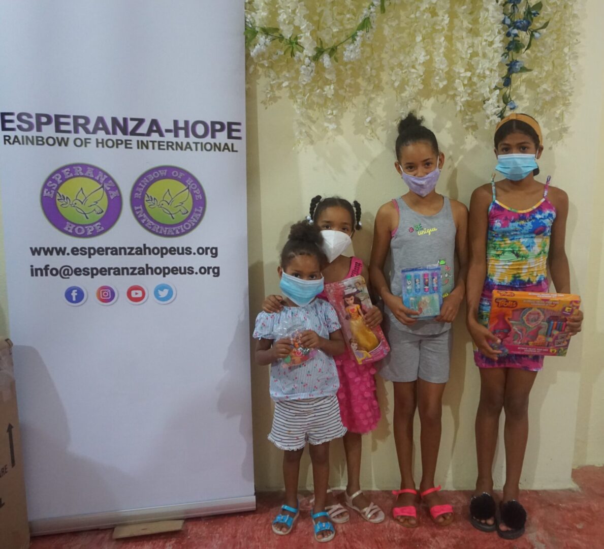 Four girls holding toys beside the Esperanza-Hope tarpaulin