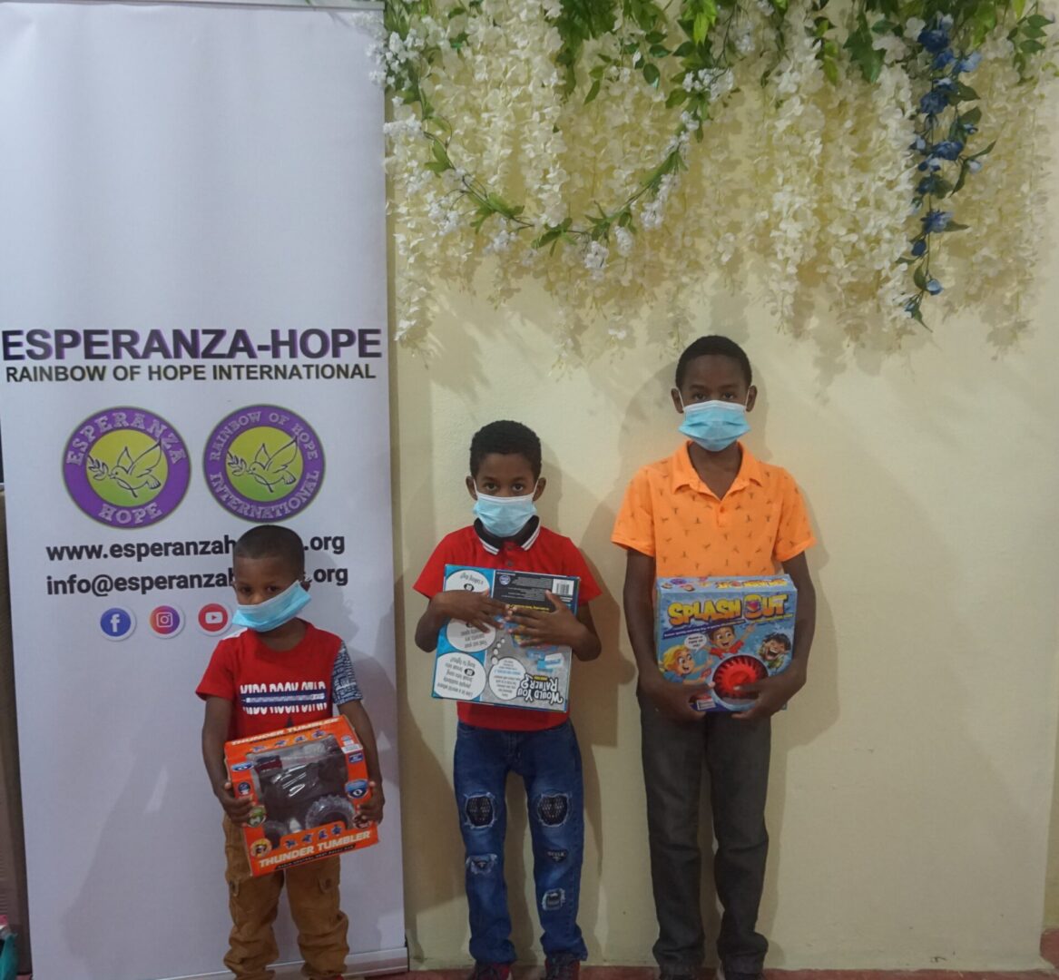 Three boys each holding a box of toy beside the Esperanza-Hope tarpaulin