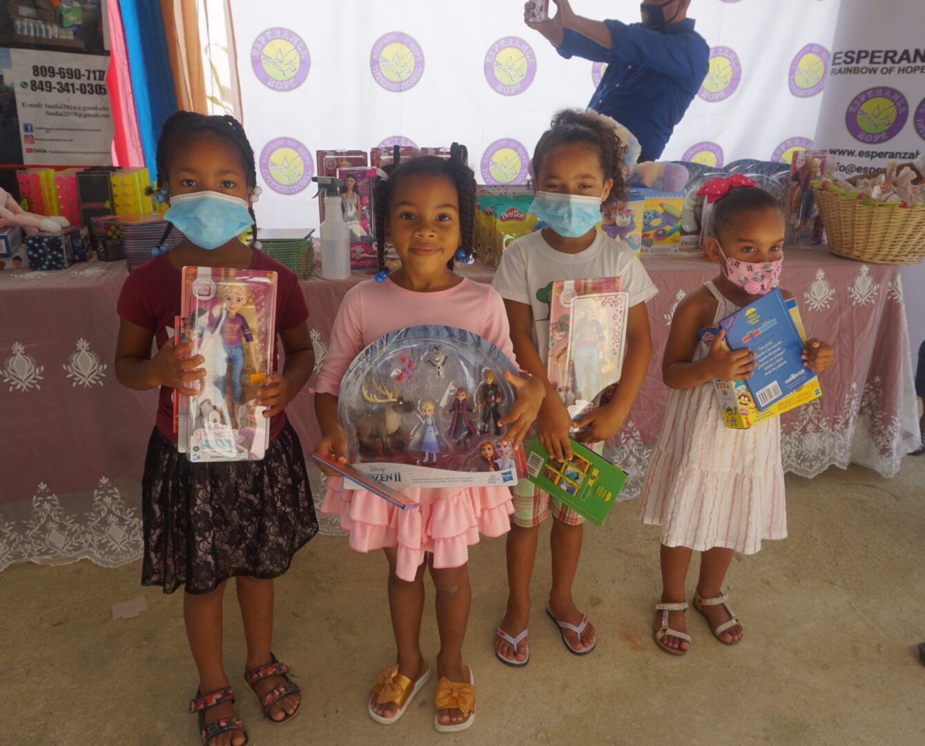 Four young girls, three wearing masks, holding books and toys