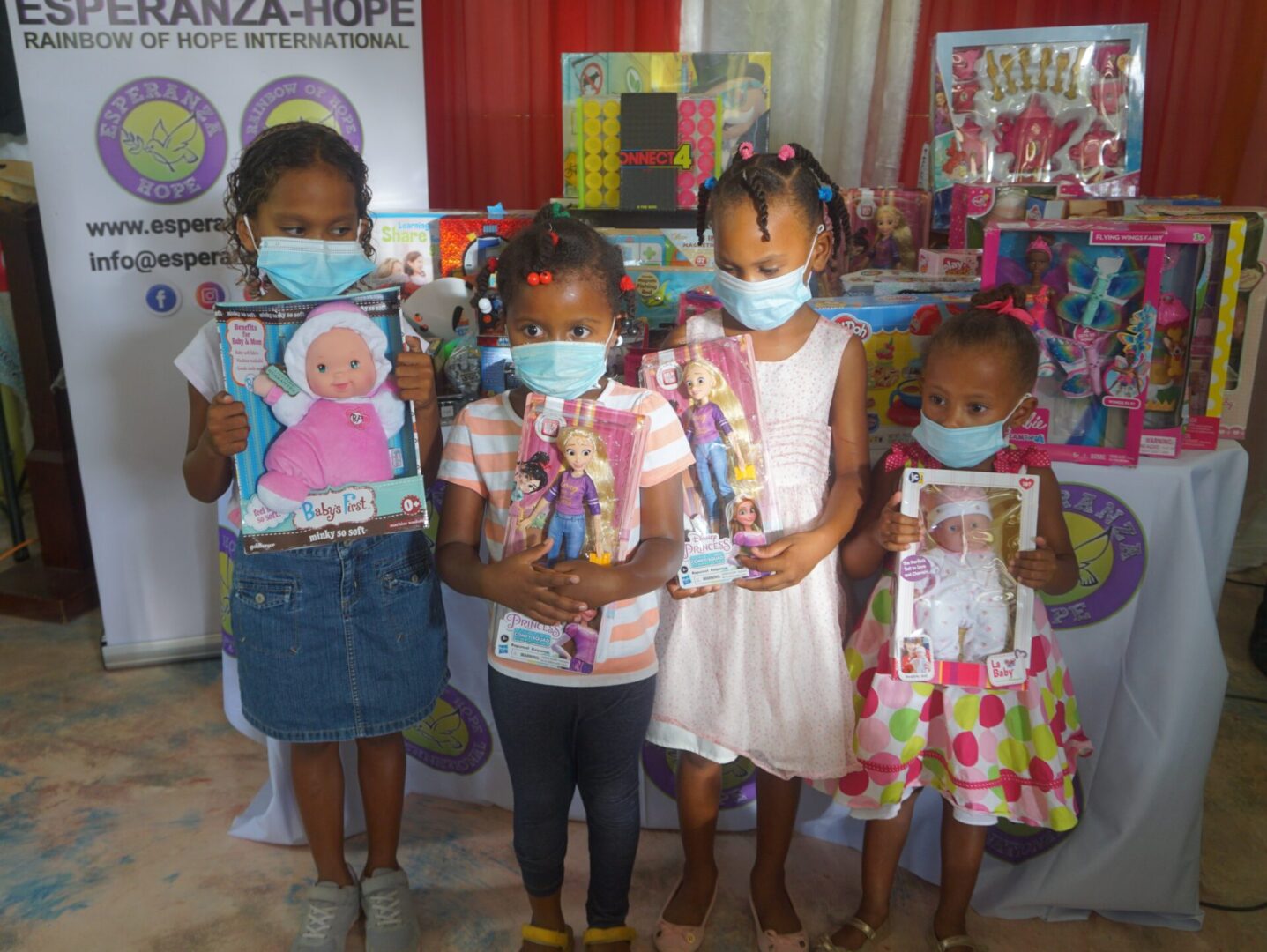 Four young girls, all wearing masks, each holding a doll in a box