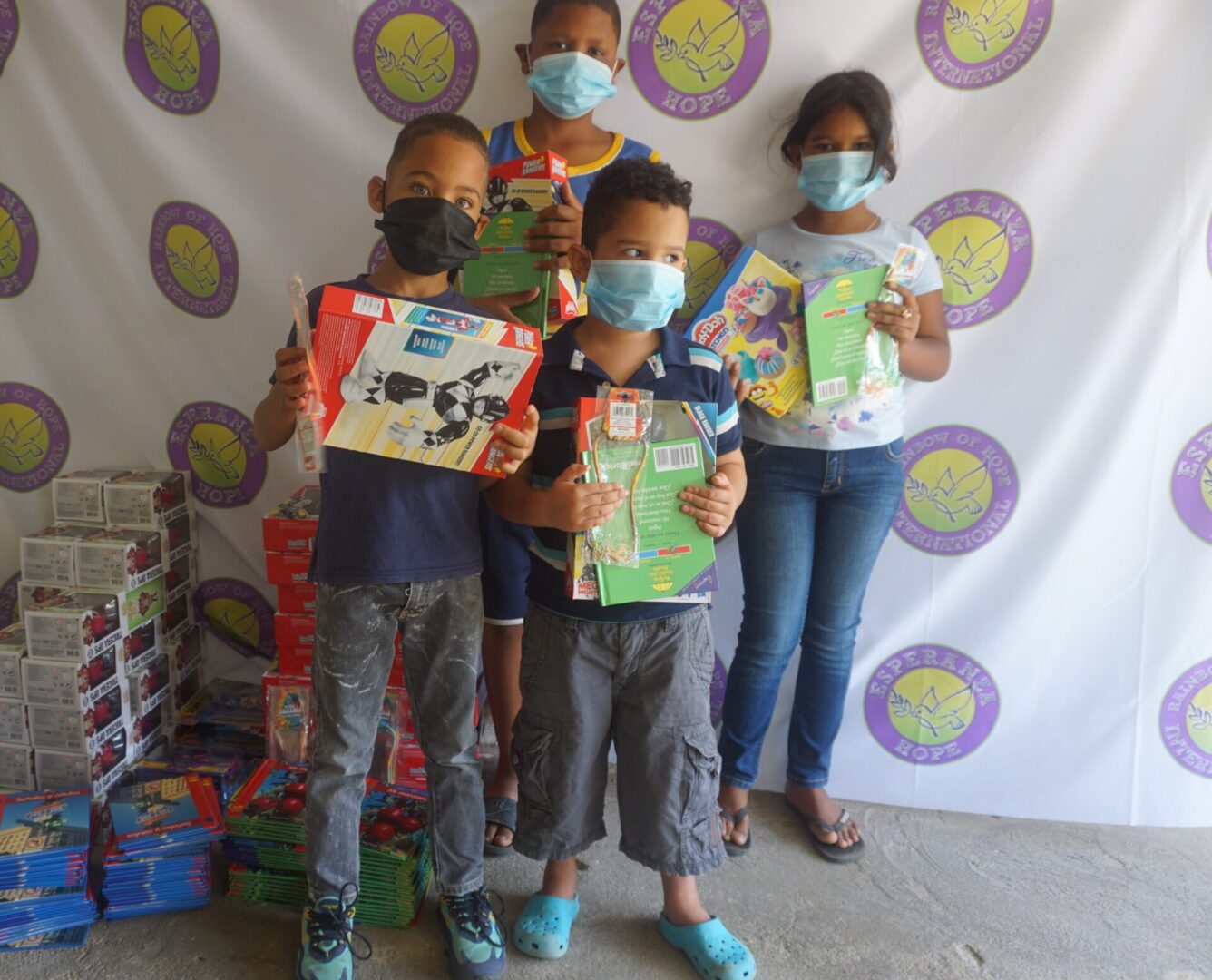 Four children standing against the Esperanza-Hope backdrop with their toys and books, batch 9